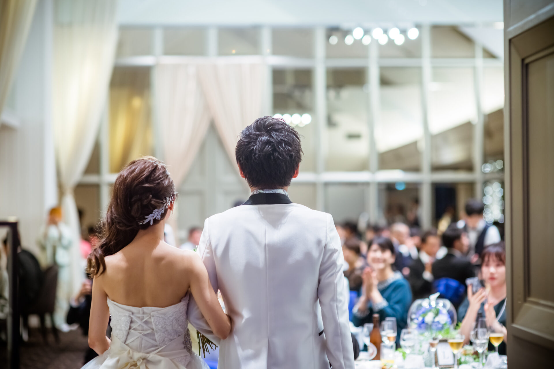 披露宴会場へ入場する新郎新婦の後ろ姿と、拍手で迎えるゲストたち